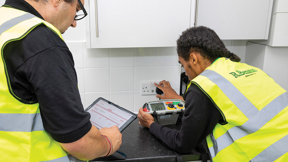 Gateway Housing EICR and remedial works - Two electricians in high-visibility vests conducting a safety test on a kitchen electrical socket. One person is using a testing device plugged into the outlet, while the other records results on a tablet.